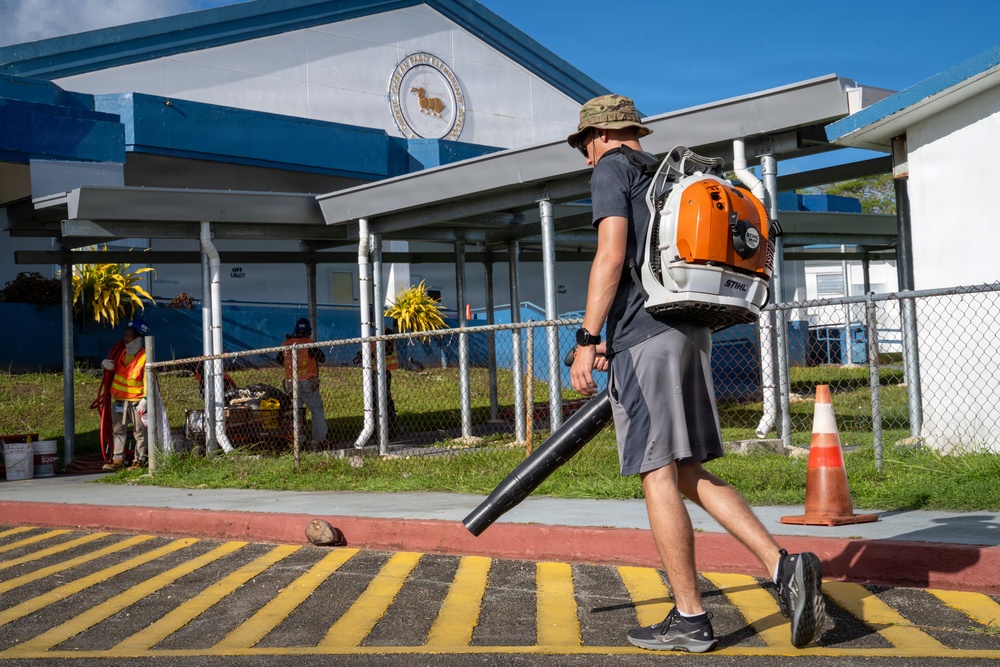 A man wearing a bucket hat and gray shorts carries a STIHL backpack leaf blower while walking across a yellow-striped speed bump or crosswalk area. The setting appears to be a school or public facility with blue and white buildings, chain-link fencing, and a decorative emblem/seal on the facade. Additional workers in safety vests are visible in the background, suggesting an organized grounds maintenance or cleanup operation.