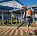 A man wearing a bucket hat and gray shorts carries a STIHL backpack leaf blower while walking across a yellow-striped speed bump or crosswalk area. The setting appears to be a school or public facility with blue and white buildings, chain-link fencing, and a decorative emblem/seal on the facade. Additional workers in safety vests are visible in the background, suggesting an organized grounds maintenance or cleanup operation.