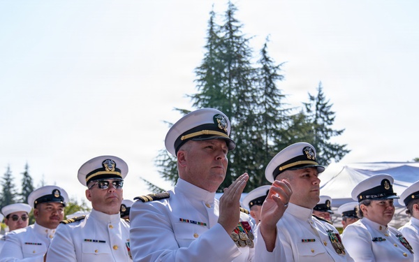 A group of U.S. Navy personnel dressed in white service dress uniforms are seated in rows of chairs at an outdoor ceremony. The individuals appear to be attending a formal military event, with several officers and enlisted members visible, some applauding. The setting is outdoors with trees and a white tent canopy visible in the background.