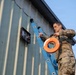 A military service member in MultiCam/OCP camouflage uniform is working on the exterior of a metal building, standing on or near a blue extension ladder. He is handling an orange fish tape spool and white cable/wire, apparently routing or pulling cable through a wall-mounted electrical or communications junction box. The work appears to be a communications or electrical installation task on a military or government facility.