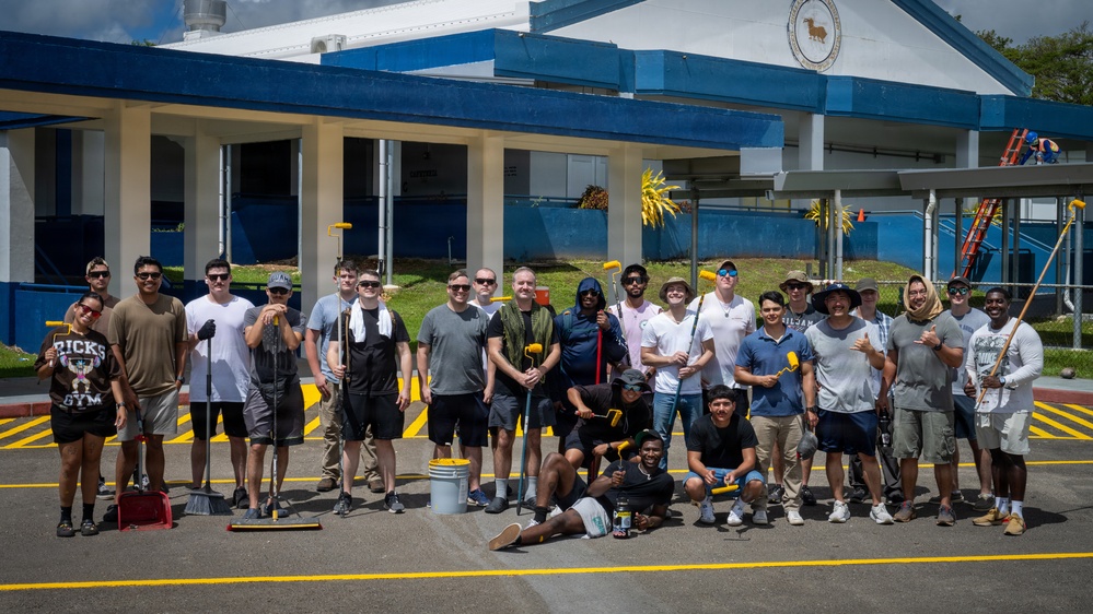 A large group of approximately 25 volunteers poses for a group photo in a parking lot after completing a community service painting project. They are holding painting tools including rollers, brushes, and brooms in front of a blue and white building. Freshly painted yellow lines are visible on the pavement, and additional workers can be seen on ladders in the background.