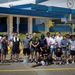 A large group of approximately 25 volunteers poses for a group photo in a parking lot after completing a community service painting project. They are holding painting tools including rollers, brushes, and brooms in front of a blue and white building. Freshly painted yellow lines are visible on the pavement, and additional workers can be seen on ladders in the background.