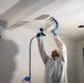 A worker in a white protective coverall suit is using an airless paint sprayer to paint or prime a ceiling in what appears to be a room under renovation. The person is wearing blue latex gloves and safety glasses while operating the spray gun with a paint cup attached. The ceiling shows signs of ongoing construction work, including an open electrical box hole and a removed ceiling panel.