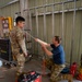 Two military personnel in camouflage uniforms are working together on a construction or installation task inside a building under renovation. One individual is standing wearing OCP (Army/Air Force) uniform with safety glasses, while the other is kneeling in what appears to be Navy NWU Type III camouflage and a blue t-shirt, both handling a metal rod or conduit. The setting shows an active worksite with metal stud framing, tools, and construction equipment visible.