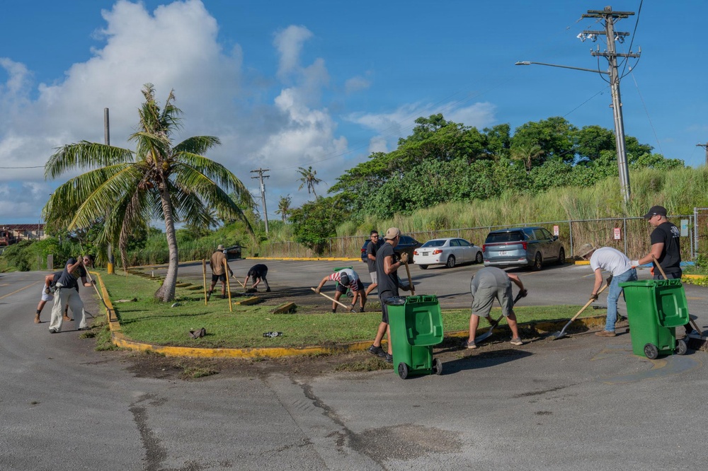 A group of approximately 8-10 people are engaged in a community cleanup or landscaping effort in a parking lot area. They are raking and sweeping debris from a grass median island surrounded by asphalt, with green waste bins positioned nearby for collection. The setting appears to be a tropical location, indicated by palm trees, lush vegetation, and a warm, sunny climate.