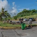 A group of approximately 8-10 people are engaged in a community cleanup or landscaping effort in a parking lot area. They are raking and sweeping debris from a grass median island surrounded by asphalt, with green waste bins positioned nearby for collection. The setting appears to be a tropical location, indicated by palm trees, lush vegetation, and a warm, sunny climate.
