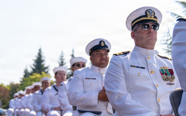 A formal U.S. Navy ceremony is taking place outdoors, with a row of naval personnel seated in folding chairs wearing white dress uniforms. The scene includes officers with medals and insignia as well as enlisted sailors, all appearing to observe a ceremony in progress. Multiple flags are visible on the left side of the frame, suggesting an official military event such as a change of command or commissioning ceremony.