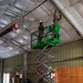 Military personnel in camouflage uniforms are performing construction or renovation work inside a large metal building. Two soldiers are elevated on a green Sunbelt scissor lift working on the ceiling, while additional personnel work at ground level. The space is filled with construction materials including rolls of insulation, plywood, and various tools.