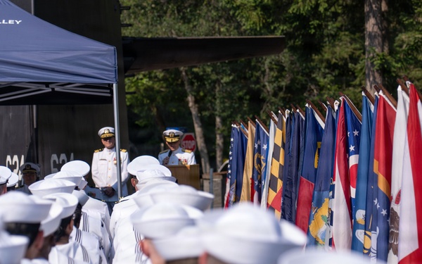 A formal U.S. Navy military ceremony is taking place outdoors, with rows of sailors in white dress uniforms standing in formation facing a podium under a canopy marked 'Trident Inn Galley.' Officers in white dress uniforms with gold insignia stand at the front addressing the assembled personnel. A large display of U.S. state flags lines the right side of the formation.