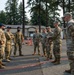 A group of military personnel in various uniforms are gathered outdoors on a paved area, appearing to be engaged in a briefing or discussion. The group includes individuals in at least two distinct uniform styles, suggesting a multinational or inter-service meeting. The setting appears to be a military installation with buildings and tall conifer trees visible in the background.