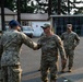 Military personnel from different branches are gathered in an outdoor area on what appears to be a military installation. Two individuals are shaking hands in the foreground, with one wearing Navy digital camouflage (NWU) and the other wearing Army/Air Force OCP (Operational Camouflage Pattern) uniform. Additional uniformed personnel are visible in the background observing the interaction.