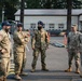 A group of military personnel from what appear to be multiple nations are gathered in a parking area at a military installation, engaged in a briefing or informal discussion. The group includes individuals wearing different camouflage patterns and headgear, including a blue turban worn by one individual, suggesting multinational participation. The setting appears to be a joint military exercise or coordination meeting at a base facility.