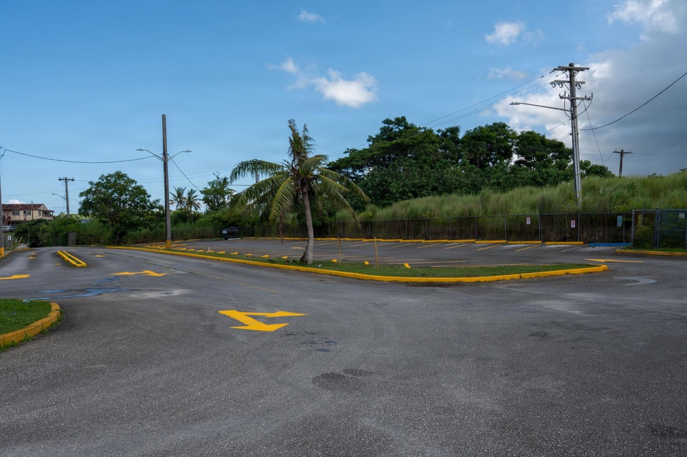 An empty parking lot with yellow-painted curbs, lane markings, and directional arrows on the asphalt surface. The lot is surrounded by tropical vegetation including a leaning palm tree, dense green trees, and tall grass behind a chain-link fence. The scene appears to be in a tropical or subtropical region, likely the Caribbean or Pacific island territory.