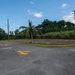 An empty parking lot with yellow-painted curbs, lane markings, and directional arrows on the asphalt surface. The lot is surrounded by tropical vegetation including a leaning palm tree, dense green trees, and tall grass behind a chain-link fence. The scene appears to be in a tropical or subtropical region, likely the Caribbean or Pacific island territory.