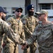 Four military personnel in camouflage uniforms are engaged in a greeting exchange outdoors, with two individuals shaking hands in the foreground. The group includes members appearing to represent different branches or units, identifiable by varying uniform patterns and insignia. One individual wears a blue Sikh dastar (turban), indicating religious accommodation within military service.