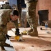 Military personnel in MultiCam/OCP camouflage uniforms are engaged in construction or facility setup work inside a building. A soldier in the foreground is using a DeWalt circular saw to cut plywood sheets while wearing safety glasses and black gloves. Multiple service members are visible in the background performing various tasks in what appears to be a construction or renovation environment.