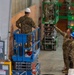 Military personnel in camouflage uniforms are engaged in construction or renovation work inside a large building. Workers are using scissor lifts — one blue and one green — to access elevated areas near walls. At least one individual on the ground is directing or supervising the work, pointing upward.