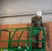A military service member wearing camouflage uniform and a white hard hat is operating a green Sunbelt scissor lift inside what appears to be a construction or renovation site. The individual is standing on the elevated platform, looking down at the lift controls. The background shows plywood wall panels and exposed insulation, indicating active construction work.