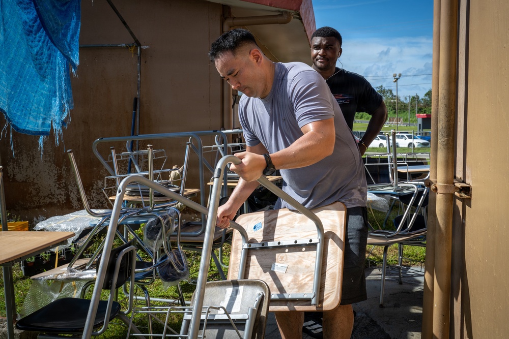 Two men are working outdoors handling metal folding chairs and furniture, likely during a cleanup, setup, or relocation effort. The foreground man in a gray t-shirt is actively gripping and moving a folding chair, while a second man in a black shirt observes from behind. The setting appears to be outside a building with various stacked chairs and tables scattered around.