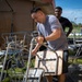 Two men are working outdoors handling metal folding chairs and furniture, likely during a cleanup, setup, or relocation effort. The foreground man in a gray t-shirt is actively gripping and moving a folding chair, while a second man in a black shirt observes from behind. The setting appears to be outside a building with various stacked chairs and tables scattered around.