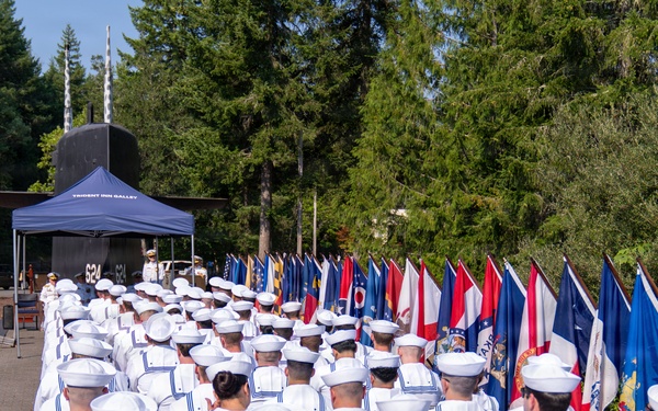 A formal U.S. Navy military ceremony is taking place outdoors, with a large formation of sailors in white dress uniforms seated in rows facing a podium or stage area. A prominent display of state and military flags lines the right side of the assembly. In the background, a submarine conning tower is visible, suggesting this event is taking place at a naval museum or memorial site.