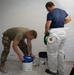 Two workers are engaged in drywall finishing or painting preparation work in what appears to be a construction or renovation space. One individual wearing military-style camouflage pants and boots is opening a bucket of joint compound, while the other in a white protective Tyvek suit holds a roll of tape and a green lid. The wall behind them shows freshly applied drywall compound and blue painter's tape markers.