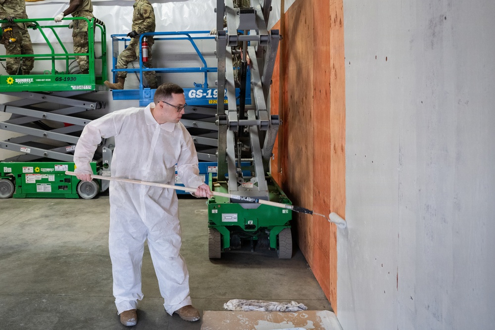 A worker in a white protective coverall suit is painting or applying a coating to a concrete wall using a long-handled roller. In the background, individuals in military camouflage uniforms are operating green scissor lift platforms branded 'Sunbelt Rentals' (model GS-1930). The setting appears to be an active construction or renovation site inside a large industrial or warehouse-type building.
