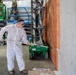 A worker in a white protective coverall suit is painting or applying a coating to a concrete wall using a long-handled roller. In the background, individuals in military camouflage uniforms are operating green scissor lift platforms branded 'Sunbelt Rentals' (model GS-1930). The setting appears to be an active construction or renovation site inside a large industrial or warehouse-type building.