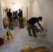 Three individuals wearing military-style camouflage uniforms and dark shirts are working inside an unfinished interior space with drywall walls and a bare concrete floor. One person is applying joint compound or spackle to a wall near electrical outlets, while two others consult near a scaffolding workbench with one pointing upward. The scene depicts active construction or renovation work with various tools and materials present.
