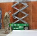 Military personnel in camouflage uniforms are engaged in construction or facility setup work inside a large building. One soldier in the foreground is using a measuring tape near a green Sunbelt Rentals scissor lift, while two others on a blue Genie GS-1930 scissor lift work on a plywood wall panel in the background. The scene suggests active interior construction or conversion of a large space.