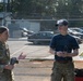 Two military personnel are engaged in conversation in an outdoor area near a chain-link fenced parking lot. The individual on the left is wearing a camouflage uniform with a name tape reading 'FOSTER' and holding a beverage container, while the person on the right wears a navy t-shirt, camouflage pants, and a baseball cap. The setting appears to be a military installation based on the uniforms, equipment visible nearby, and the secured compound in the background.