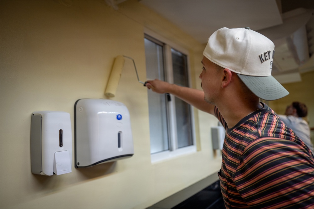 A young man wearing a white baseball cap and striped shirt is painting a wall using a paint roller near a window. The wall features mounted dispensers, likely soap and paper towel dispensers, suggesting an institutional or commercial setting such as a school, office, or community facility. The scene appears to be a renovation or maintenance activity.