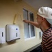 A young man wearing a white baseball cap and striped shirt is painting a wall using a paint roller near a window. The wall features mounted dispensers, likely soap and paper towel dispensers, suggesting an institutional or commercial setting such as a school, office, or community facility. The scene appears to be a renovation or maintenance activity.