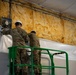Two individuals in military camouflage uniforms and white hard hats are working on an elevated green scissor lift platform. They appear to be installing or adjusting a ventilation fan unit on a wall, with exposed fiberglass batt insulation visible along the upper wall section. The setting appears to be an interior construction or facility setup environment.