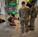 A group of military personnel in OCP (Operational Camouflage Pattern) uniforms are engaged in a construction or electrical work task inside a building. One service member is kneeling on the floor operating a power tool, while others observe and assist. A Sunbelt Rentals scissor lift (model GS-1930) is visible in the background with additional personnel on it.