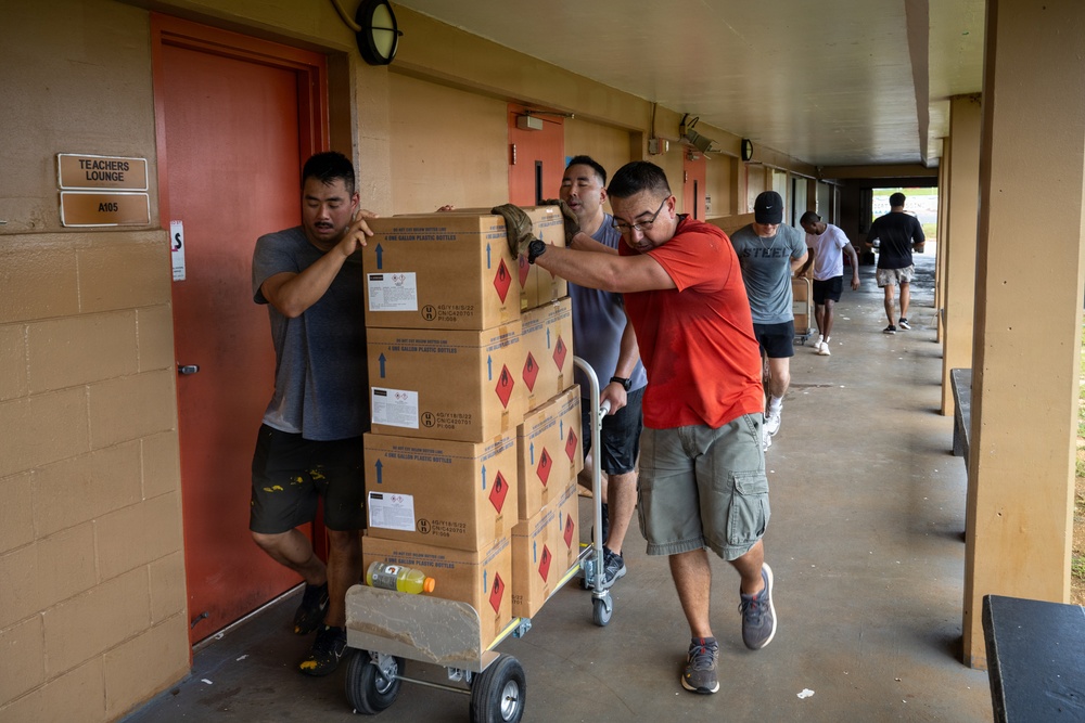 A group of people are moving stacked cardboard boxes on a hand truck along an outdoor covered corridor of what appears to be a school building. The boxes bear red diamond hazard warning symbols, indicating flammable or hazardous contents. Multiple individuals are working together to push the heavily loaded dolly toward a room marked 'Teachers Lounge A105'.