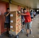 A group of people are moving stacked cardboard boxes on a hand truck along an outdoor covered corridor of what appears to be a school building. The boxes bear red diamond hazard warning symbols, indicating flammable or hazardous contents. Multiple individuals are working together to push the heavily loaded dolly toward a room marked 'Teachers Lounge A105'.