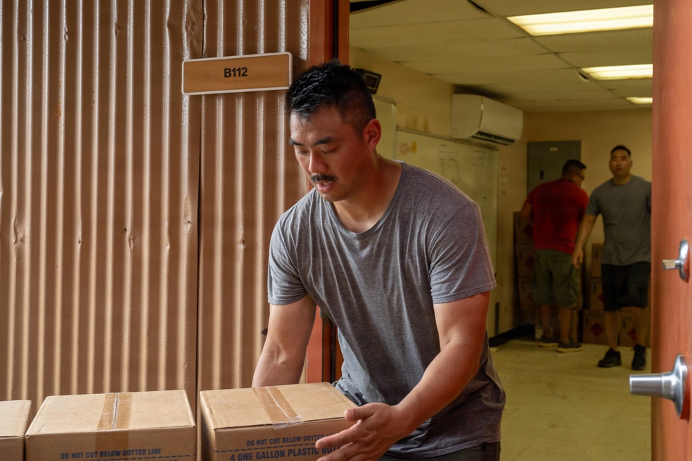 A man in a gray t-shirt is handling cardboard boxes in what appears to be a storage or warehouse corridor. The boxes are labeled '4 ONE GALLON PLASTIC' and marked 'DO NOT CUT BELOW DOTTED LINE.' Two other individuals are visible in the background of the hallway, also engaged in moving or organizing items.