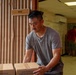 A man in a gray t-shirt is handling cardboard boxes in what appears to be a storage or warehouse corridor. The boxes are labeled '4 ONE GALLON PLASTIC' and marked 'DO NOT CUT BELOW DOTTED LINE.' Two other individuals are visible in the background of the hallway, also engaged in moving or organizing items.