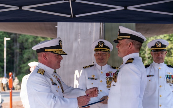 A formal U.S. Navy military ceremony is taking place outdoors under a dark canopy tent. Senior naval officers in white dress uniforms are engaged in what appears to be an award presentation or change of command ceremony, with one officer presenting a document or award to another. The setting includes arranged chairs suggesting an audience was present for the formal occasion.