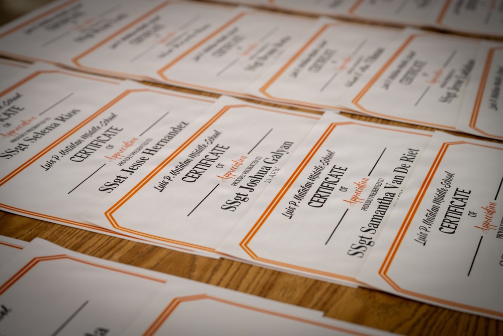 Multiple Certificates of Appreciation from Luis P. Matidan Middle School are spread across a wooden table. The certificates are awarded to military staff sergeants (SSgt), with visible names including Joshua Galvan, Jesse Hernandez, Selena Rios, Samantha Van De Riet, and others. The certificates feature an orange and black border design on white paper.