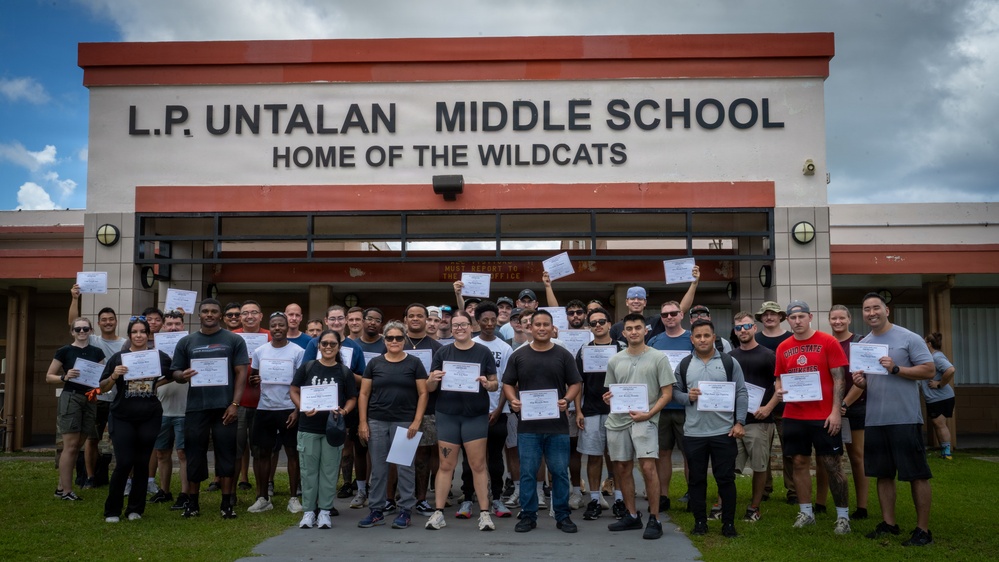 A large group of approximately 40 adults poses in front of L.P. Untalan Middle School, 'Home of the Wildcats,' holding certificates. The group appears to have completed a training or certification program, as evidenced by the documents they are proudly displaying. The photo appears to be an official commemorative group shot taken outdoors under partly cloudy skies.