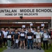 A large group of approximately 40 adults poses in front of L.P. Untalan Middle School, 'Home of the Wildcats,' holding certificates. The group appears to have completed a training or certification program, as evidenced by the documents they are proudly displaying. The photo appears to be an official commemorative group shot taken outdoors under partly cloudy skies.