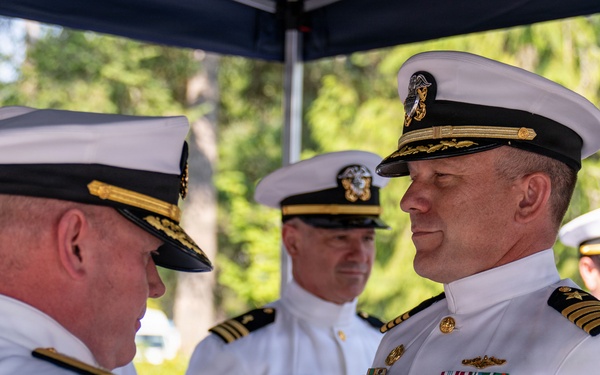 A formal U.S. Navy military ceremony is taking place outdoors, in which one officer is pinning a medal or decoration onto another officer's uniform. Both primary figures are wearing white dress uniforms with officer caps, and the recipient displays multiple medals on his chest. Additional naval officers are visible in the background observing the ceremony.