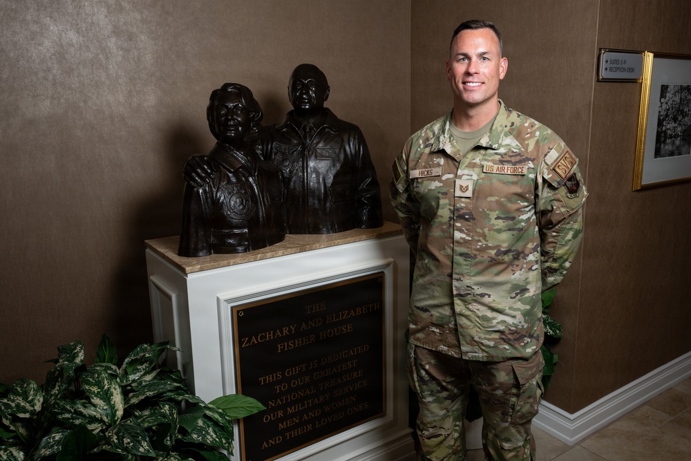 A U.S. Air Force service member identified by name tape as 'Hicks' stands beside a bronze bust memorial display at a Fisher House facility. The soldier is wearing OCP (Operational Camouflage Pattern) uniform with U.S. Air Force tape and rank insignia visible. The setting appears to be an indoor lobby or reception area of a Fisher House, a charitable organization supporting military families.