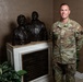 A U.S. Air Force service member identified by name tape as 'Hicks' stands beside a bronze bust memorial display at a Fisher House facility. The soldier is wearing OCP (Operational Camouflage Pattern) uniform with U.S. Air Force tape and rank insignia visible. The setting appears to be an indoor lobby or reception area of a Fisher House, a charitable organization supporting military families.