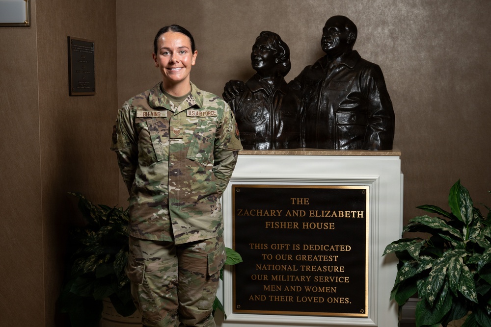 A U.S. Air Force service member wearing OCP (Operational Camouflage Pattern) uniform with the name tape 'BLEVINS' stands beside a dedication plaque and bronze sculpture at a Fisher House location. The plaque reads 'The Zachary and Elizabeth Fisher House' with a dedication to military service men and women and their loved ones. The setting appears to be an interior lobby or entrance area of a Fisher House facility.