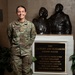 A U.S. Air Force service member wearing OCP (Operational Camouflage Pattern) uniform with the name tape 'BLEVINS' stands beside a dedication plaque and bronze sculpture at a Fisher House location. The plaque reads 'The Zachary and Elizabeth Fisher House' with a dedication to military service men and women and their loved ones. The setting appears to be an interior lobby or entrance area of a Fisher House facility.