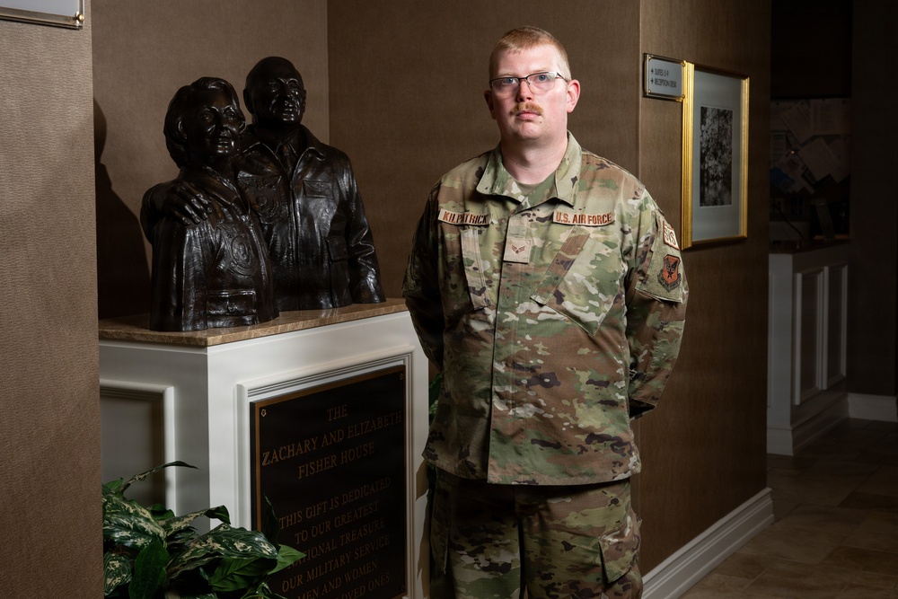 A U.S. Air Force service member in OCP (Operational Camouflage Pattern) uniform stands beside a bronze bust sculpture and dedication plaque at the Zachary and Elizabeth Fisher House. The individual wears glasses and has a name tape reading 'KILPATRICK' on their uniform. The setting appears to be an interior hallway or lobby of a Fisher House facility, which provides housing for military families.