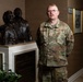 A U.S. Air Force service member in OCP (Operational Camouflage Pattern) uniform stands beside a bronze bust sculpture and dedication plaque at the Zachary and Elizabeth Fisher House. The individual wears glasses and has a name tape reading 'KILPATRICK' on their uniform. The setting appears to be an interior hallway or lobby of a Fisher House facility, which provides housing for military families.
